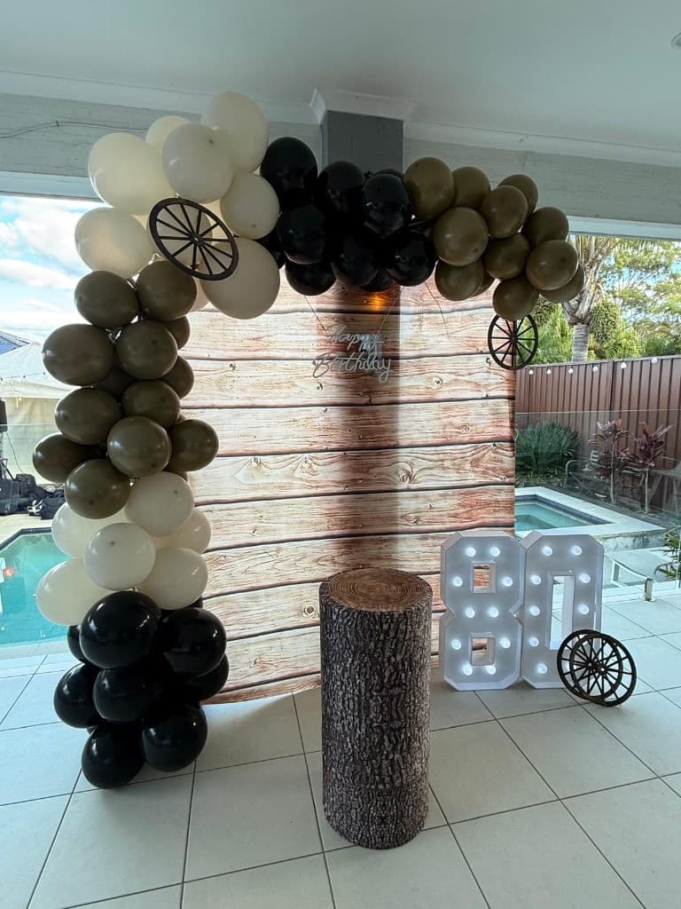 80th birthday styling with wood backdrop, balloon arch, light-up numbers, and tree stump plinth